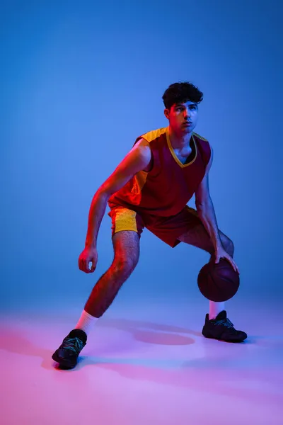 Studio shot młodego człowieka, profesjonalny koszykarz gra w koszykówkę odizolowany na niebieskim tle w neonowym świetle. — Zdjęcie stockowe