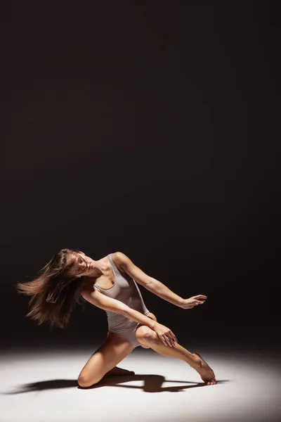 Ritratto dinamico di una giovane ballerina flessibile che danza isolata su uno sfondo scuro da studio sotto i riflettori. Arte, bellezza, concetto di ispirazione. — Foto Stock
