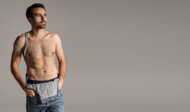 Gri stüdyo arka planında izole edilmiş, kot pantolonlu, sakalsız genç bir adamın portresi..