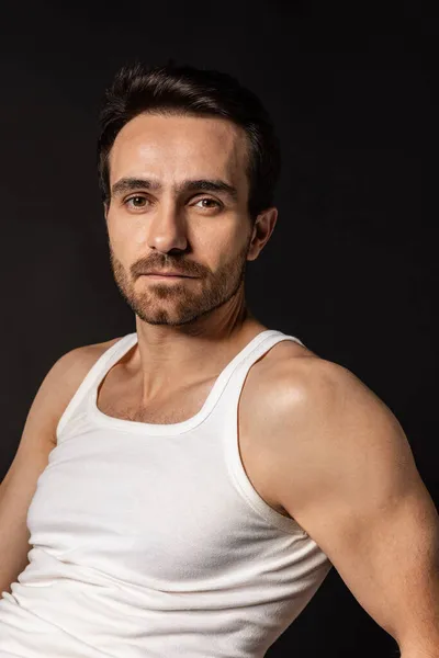 Retrato de primer plano de un joven guapo barbudo con una camiseta blanca aislada sobre fondo oscuro del estudio. Emociones, expresión facial — Foto de Stock
