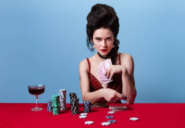 Koyu kırmızı, şarap rengi gece elbisesi giyen genç ve güzel bir kadın bahse girer, mavi arka planda poker oynar..