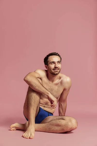 Studio laukaus nuori komea parrakas parraton mies sinisissä pikkuhousuissa istuu lattialla eristetty vaaleanpunainen tausta. — kuvapankkivalokuva