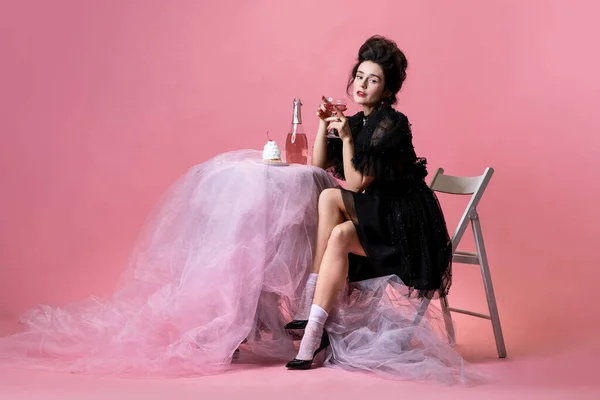 Νεαρή όμορφη γυναίκα με ρετρό στυλ χτένισμα σε μαύρο βράδυ μικρό φόρεμα κάθεται σε τραπέζι διακοπών απομονωμένο σε ροζ φόντο. — Φωτογραφία Αρχείου