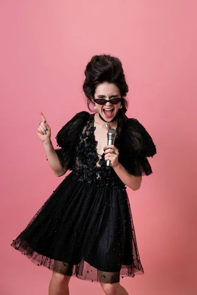 Retrato creativo de una joven adorable en vestido de noche negro cantando aislado sobre fondo rosa. Concepto de vacaciones, moda, estilo — Foto de Stock