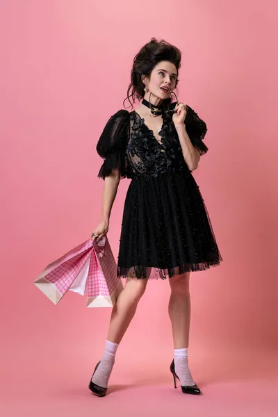 Portret młodej uroczej dziewczyny w czarnej sukni wieczorowej z torbami na zakupy odizolowanymi na różowym tle. Pojęcie sprzedaży, mody, stylu — Zdjęcie stockowe
