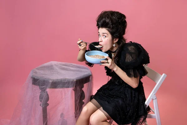 Mulher bonita nova com penteado estilo retro em preto vestido pequeno noite sentado à mesa e comer mingau isolado no fundo rosa. — Fotografia de Stock