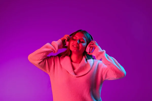 Menina bonita sorridente nova ouvindo música em fones de ouvido isolados no fundo do estúdio roxo no filtro rosa néon. Conceito de emoções, — Fotografia de Stock