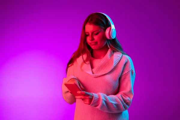 Genç, gülümseyen güzel bir kız kulaklıkla müzik dinliyor. Mor stüdyo arka planında, neon pembe filtreyle izole edilmiş. Duyguların kavramı, — Stok fotoğraf