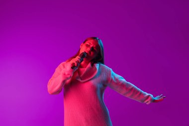 Genç, gülümseyen, mikrofonla şarkı söyleyen güzel bir kız. Mor stüdyo arka planında, neon pembe filtreyle izole edilmiş. Duyguların kavramı,