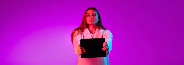 Ein junges glückliches Mädchen in warmem Strickpullover mit Tablet, Gerät isoliert über lila Studiohintergrund in Neonlicht. — Stockfoto