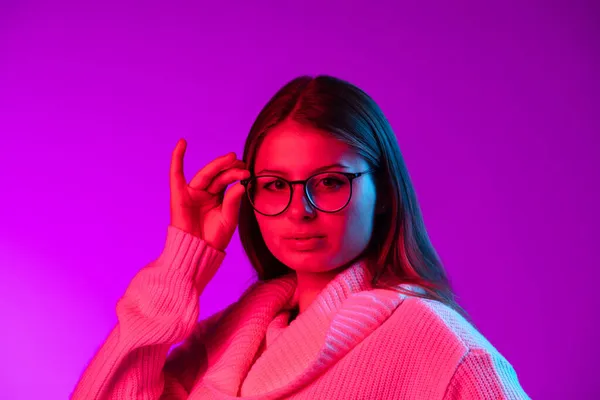 Retrato de chica bonita joven en suéter de punto caliente poniendo gafas en aislado sobre fondo de estudio púrpura en luz de neón. — Foto de Stock