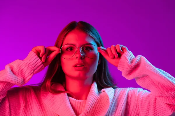 Potret gadis muda cantik dengan sweater rajutan hangat menempatkan kacamata di terisolasi atas latar belakang studio ungu di lampu neon. — Stok Foto