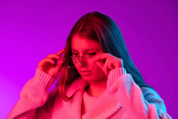 Portrait of young pretty girl in warm knitted sweater putting eyeglasses on isolated over purple studio background in neon light. — Stock Photo, Image
