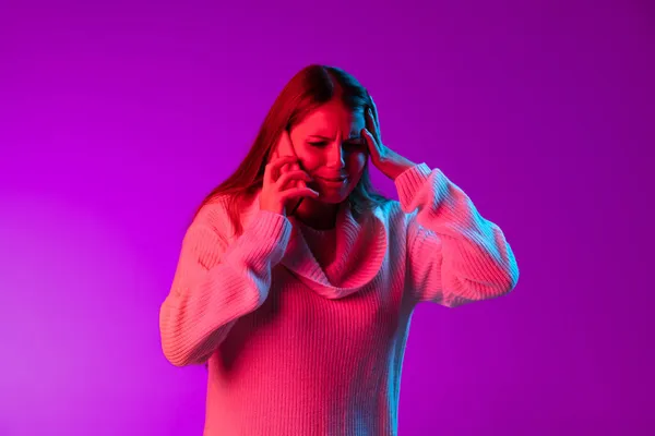 Retrato de menina bonita jovem em suéter de malha quente usando telefone isolado sobre fundo estúdio roxo em luz de néon. — Fotografia de Stock