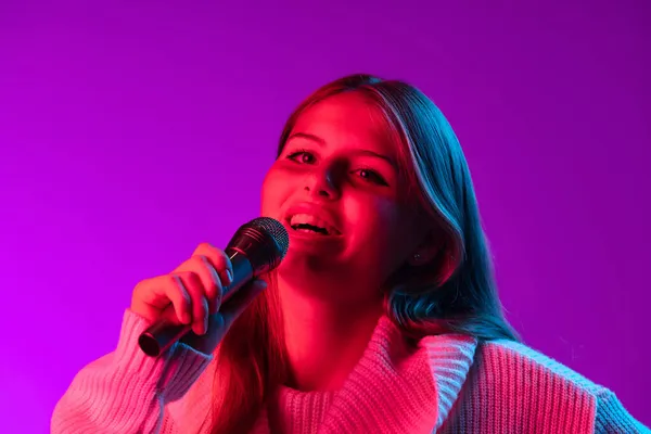 Close-up jong glimlachend mooi meisje zingen met microfoon geïsoleerd op paarse studio achtergrond in neon roze filter. Concept van emoties, — Stockfoto