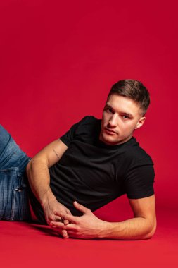 Siyah tişörtlü, mavi kotlu yakışıklı bir adamın portresi kırmızı stüdyo arka planında izole bir şekilde yerde yatıyor..