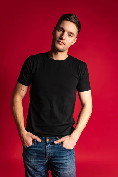 Muotokuva nuori komea urheilullinen mies musta t-paita ja sininen farkut poseeraa eristetty punainen studio tausta. — kuvapankkivalokuva