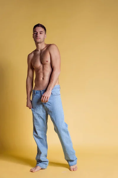 Ritratto di giovane bell'uomo senza camicia in jeans blu in posa isolato su sfondo giallo studio. — Foto Stock