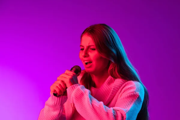 Jovem sorrindo menina bonita cantando com microfone isolado no fundo do estúdio roxo em neon rosa filtro. Conceito de emoções, — Fotografia de Stock