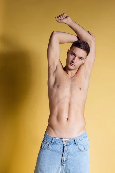 Retrato de mitad de longitud de un joven guapo y deportivo hombre sin camisa en pantalones vaqueros azules de pie con las manos arriba aisladas sobre un fondo amarillo. —  Fotos de Stock