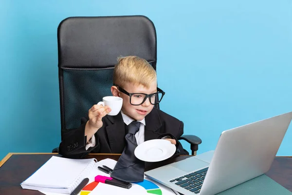Porträtt av liten pojke, barn affärsman i enorm jacka kostym sitter vid kontorsbordet isolerad på blå studio bakgrund. — Stockfoto
