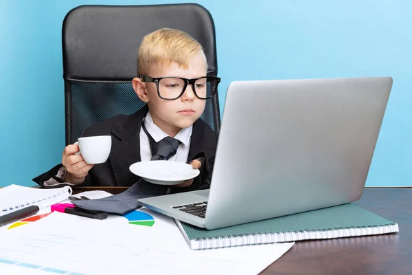 Ritratto di bambino, uomo d'affari bambino in giacca enorme vestito seduto al tavolo dell'ufficio e utilizzando computer portatile isolato su sfondo blu studio. — Foto Stock