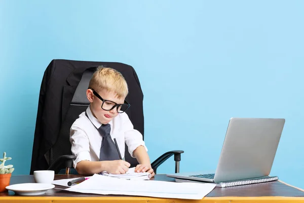 Een kleine jongen, een kinderzakenman zit aan een kantoortafel geïsoleerd op een blauwe studioachtergrond. Concept van slimme kindertijd — Stockfoto