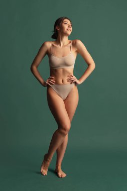 Çıplak renkli iç çamaşırlı, koyu yeşil stüdyo arka planında izole edilmiş genç ve zayıf bir kadının portresi. Doğal güzellik konsepti.
