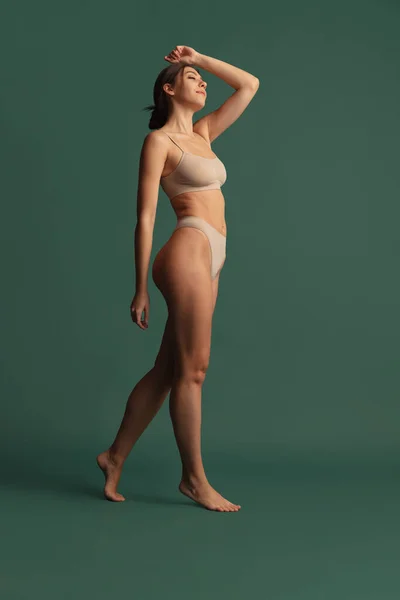 Retrato de jovem bela mulher magra em roupa interior de cor nua posando isolado sobre fundo estúdio verde escuro. Conceito de beleza natural. — Fotografia de Stock