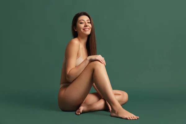 Νεαρό όμορφο χαμογελαστό κορίτσι σε γυμνό χρώμα εσώρουχα κάθεται στο πάτωμα απομονωμένο πάνω από σκούρο πράσινο φόντο στούντιο. Έννοια φυσικής ομορφιάς. — Φωτογραφία Αρχείου