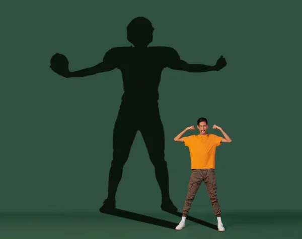Conceptual image with young boy dreaming about future sport career and shadow of fit male football player on dark green background — Stock Photo, Image