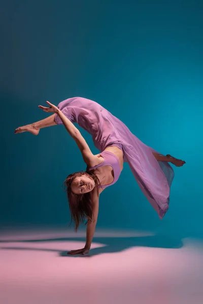 Молодой очаровательный гибкий танцор в сиреневом платье танца изолированы на градиентно-голубом фоне в неоновом. — стоковое фото