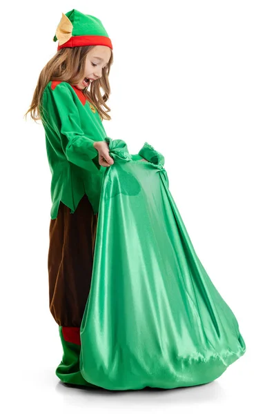 Здивована маленька дівчинка, одягнена як смішний гном або ельф з великою зеленою подарунковою сумкою, ізольована на білому фоні студії. Зима, свято, різдвяна концепція — стокове фото