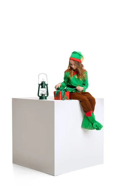 Маленькая красивая девочка, одетая как смешной гном или эльф, сидящая на большой коробке, изолированной на белом фоне студии. Зима, праздник, рождественская концепция — стоковое фото