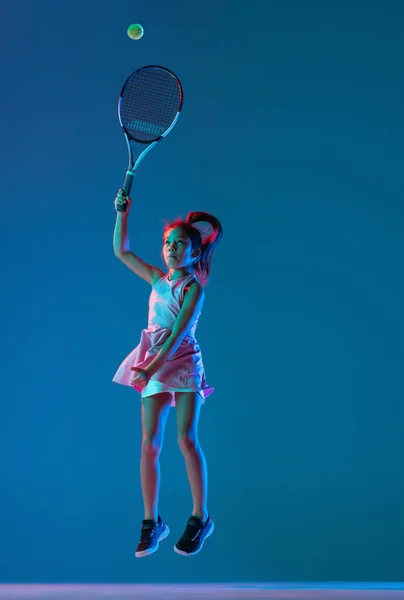 Porträt eines kleinen Mädchens, Tennisanfängerin, die isoliert auf blauem Studiohintergrund in Neonlicht Tennis spielt. Sport, Studium, Kindheitskonzept — Stockfoto