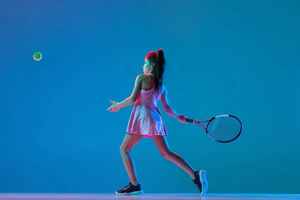 Estúdio tiro de menina, iniciante treinamento de tenista isolado no fundo do estúdio azul em luz de néon. Esporte, estudo, conceito de infância — Fotografia de Stock