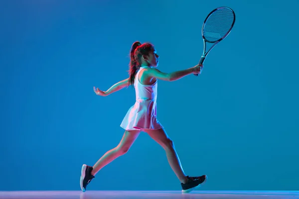 Студійний знімок маленької дівчинки, тренування тенісиста-початківця ізольовано на синьому студійному фоні в неоновому світлі. Спорт, навчання, концепція дитинства — стокове фото