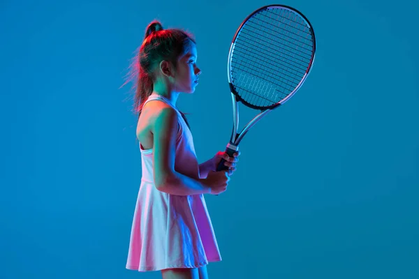 Vista de perfil de niña, tenista principiante posando con raqueta aislada sobre fondo azul estudio en luz de neón. Deporte, estudio, concepto infantil —  Fotos de Stock