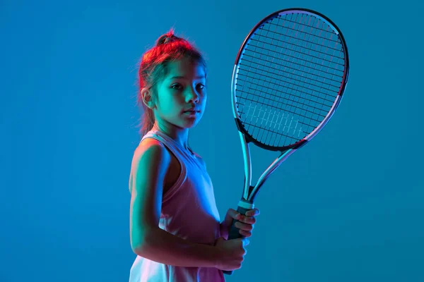Pół-długości portret dziewczynki, początkujący tenisista pozujący z rakietą odizolowaną na niebieskim tle studia w neonowym świetle. Sport, nauka, koncepcja dzieciństwa — Zdjęcie stockowe