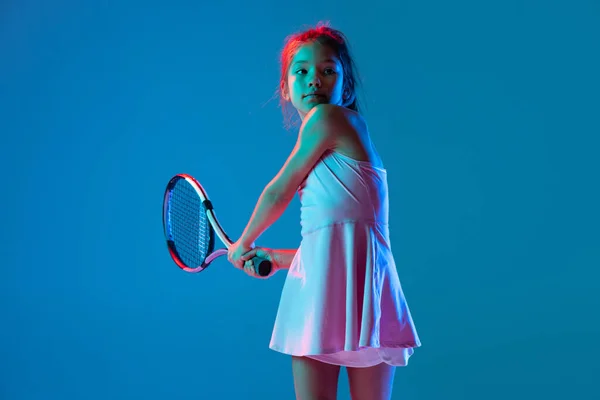 Portret dziewczynki, początkujący tenisista grający w tenisa na niebieskim tle studia w neonowym świetle. Sport, nauka, koncepcja dzieciństwa — Zdjęcie stockowe