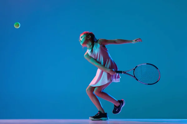 Studio skott av liten flicka, nybörjare tennisspelare utbildning isolerad på blå studio bakgrund i neon ljus. Idrott, studier, barndomskoncept — Stockfoto