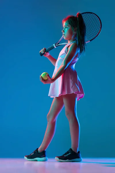 Een klein meisje, beginnende tennisser die geïsoleerd poseert op een blauwe studio achtergrond in neon licht. Sport, studie, jeugdconcept — Stockfoto