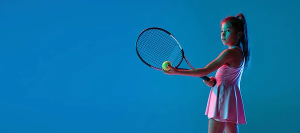 Studyjne ujęcie małej dziewczynki, początkujący tenisista trening izolowany na niebieskim tle studio w neonowym świetle. Sport, nauka, koncepcja dzieciństwa — Zdjęcie stockowe