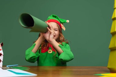 Komik cüce gibi giyinmiş mutlu çocuk portresi ya da masada oturan ve Noel Baba 'ya yeşil arka planda izole edilmiş mektuplar yazan elf..