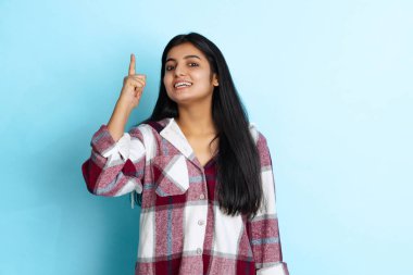 Genç gülümseyen kız portresi, sıcak kareli gömlekli öğrenci açık mavi stüdyo arka planında izole edilmiş.