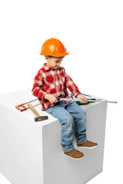 Menino pré-escolar bonito, criança na imagem de construtor, arquiteto em capacete protetor laranja sentado em enorme caixa isolada no fundo branco — Fotografia de Stock