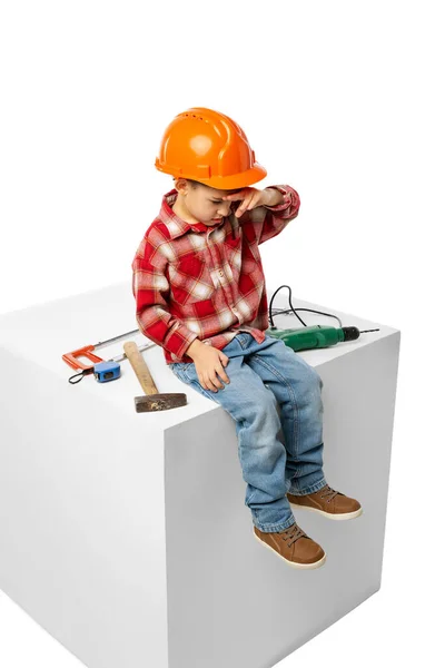 Lindo niño preescolar, niño en la imagen del constructor, arquitecto en casco protector naranja sentado en una caja enorme aislado sobre fondo blanco — Foto de Stock