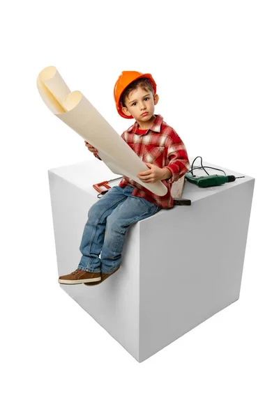 Ute pequeño niño preescolar, niño en la imagen del constructor, arquitecto en casco protector naranja sentado en una caja enorme aislado sobre fondo blanco — Foto de Stock