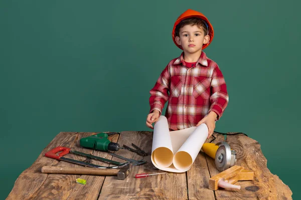Porträtt av söt liten pojke, unge i bild av byggmästare, designer i orange skyddshjälm med hjälp av arbetsverktyg isolerade på grön bakgrund — Stockfoto