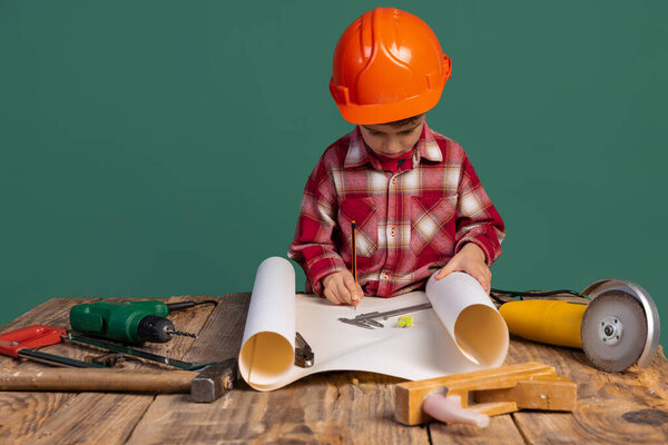 Portrait of cute little boy, kid in image of builder, designer in orange protective helmet using work tools isolated on green background